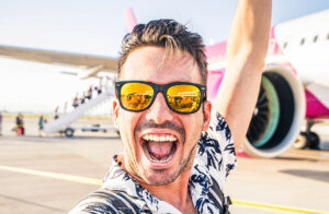 fotografia de um homem branco com óculos escuros com expressão de felicidade e um avião ao fundo